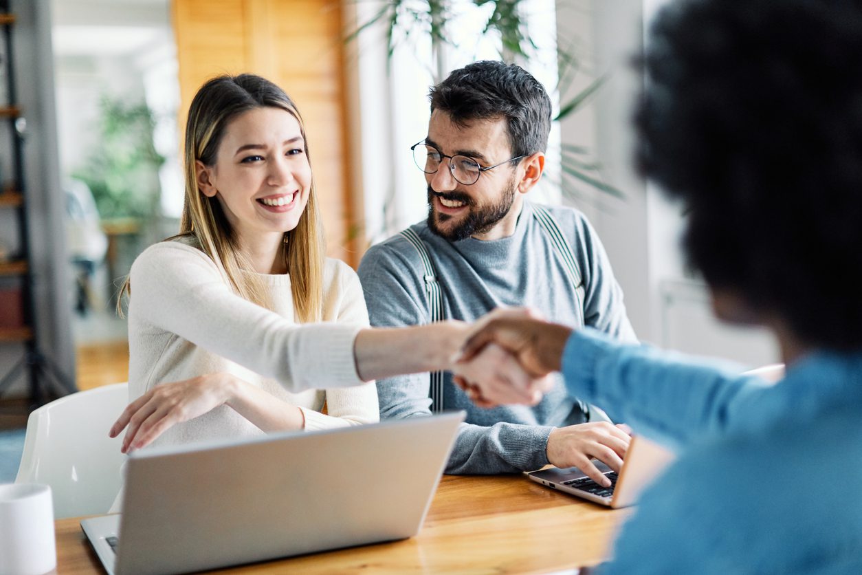 A man and woman across a table from another woman. The man and woman on one side of the table each have a laptop. The two women are shaking hands