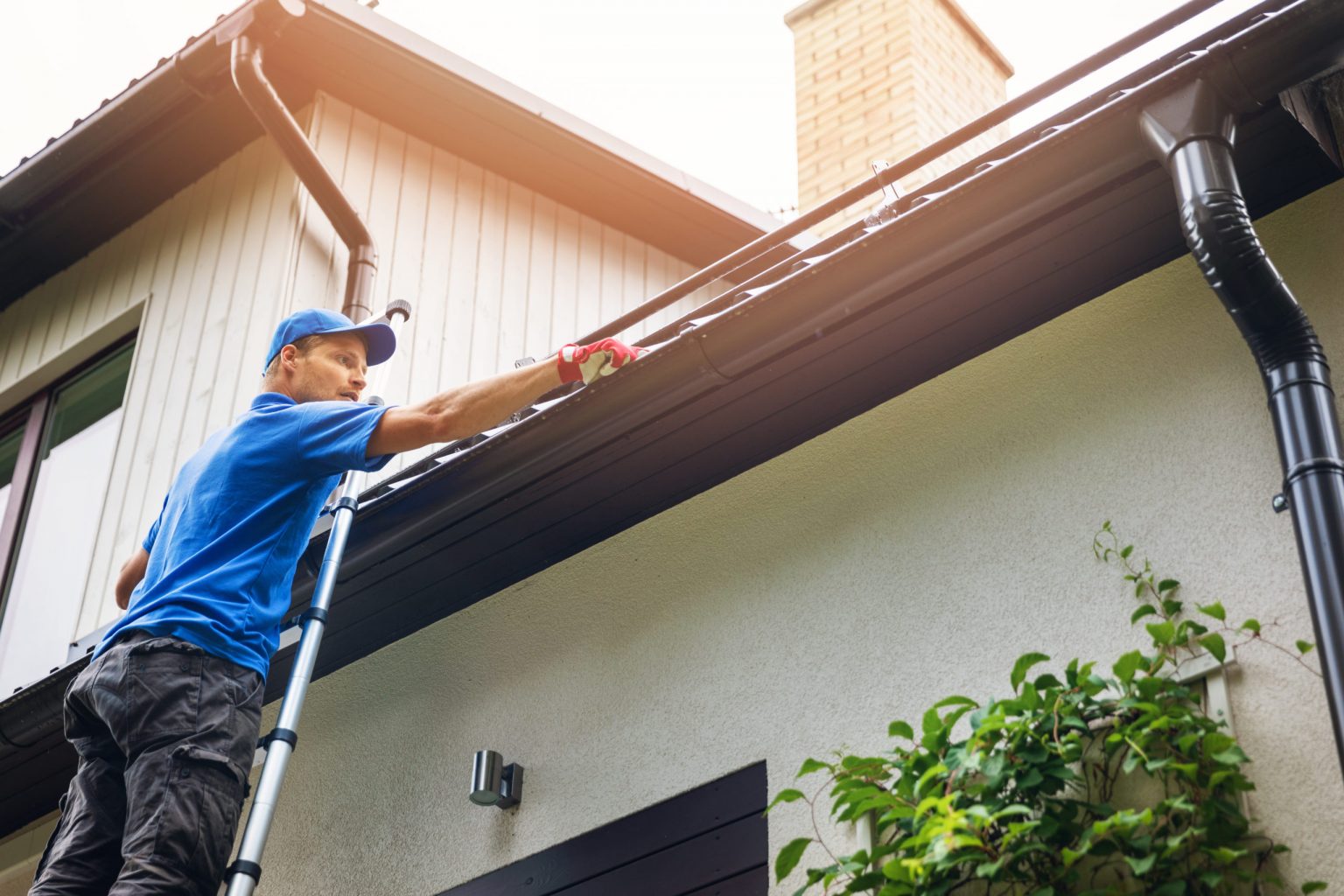 A man wearing a blue shirt and blue baseball cap cleaning the gutters of a two-story home