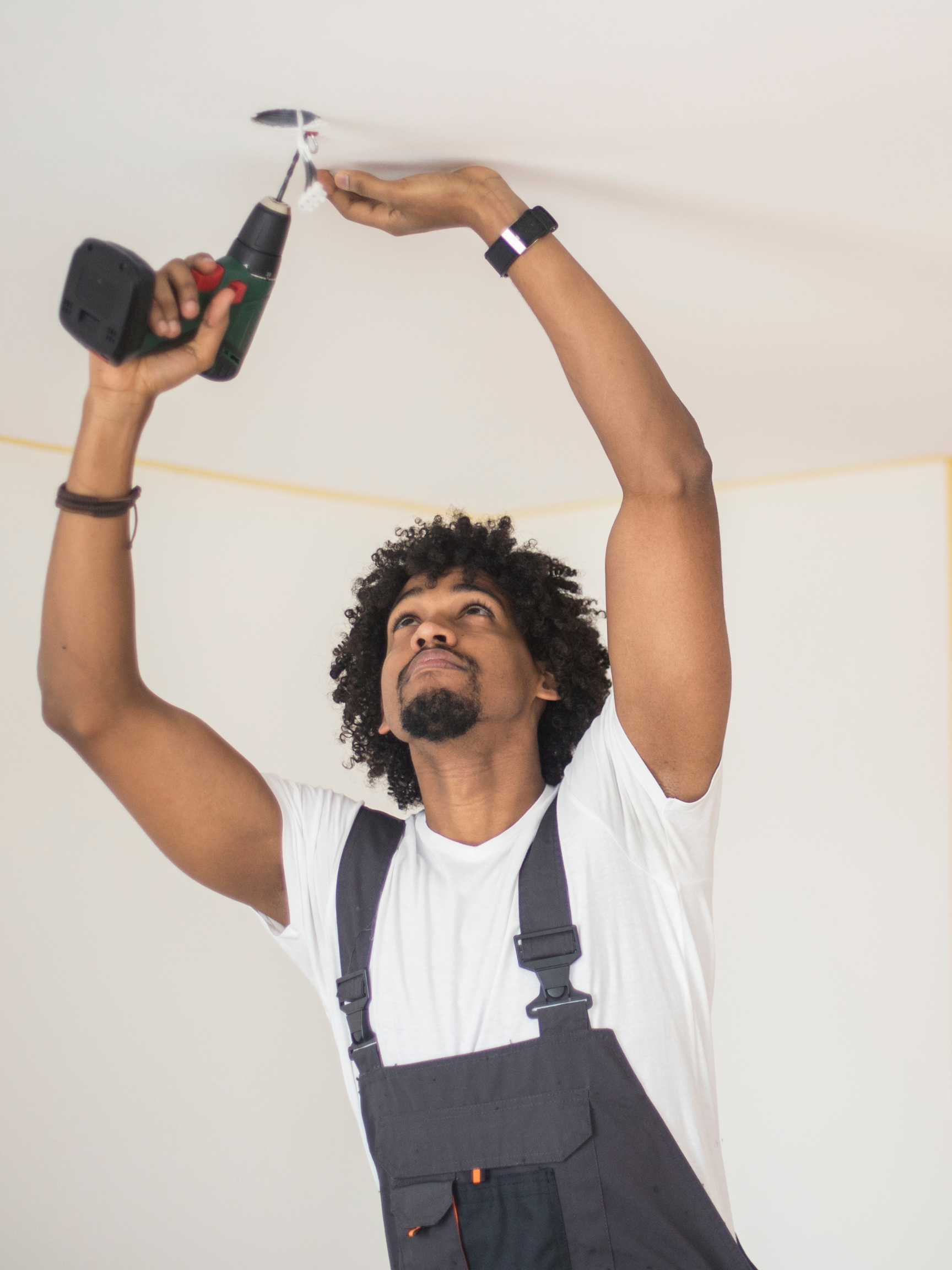 A man waring overalls and a white t-shirt using a drill to work on the electrical wiring in the ceiling